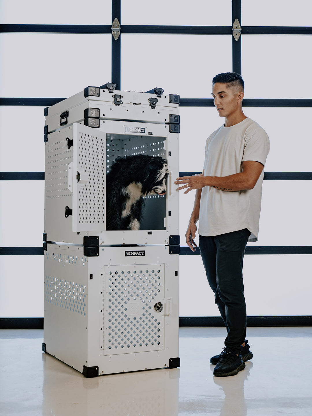 man standing next to three crates stacked with a dog in the crate. The crates are stacked, white stationary crate, white high anxiety dog crate with a fluffy black and white dog inside of it, and the top crate is a collapsed collapsible dog crate that is also white